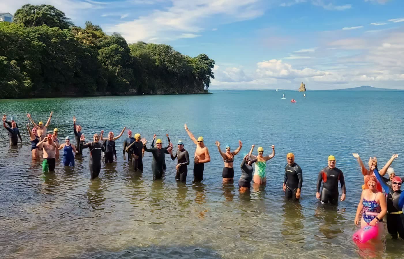 Hibiscus Coast Triathlon Club members in the water after a group swim, arms raised, with Rangitoto in the distance